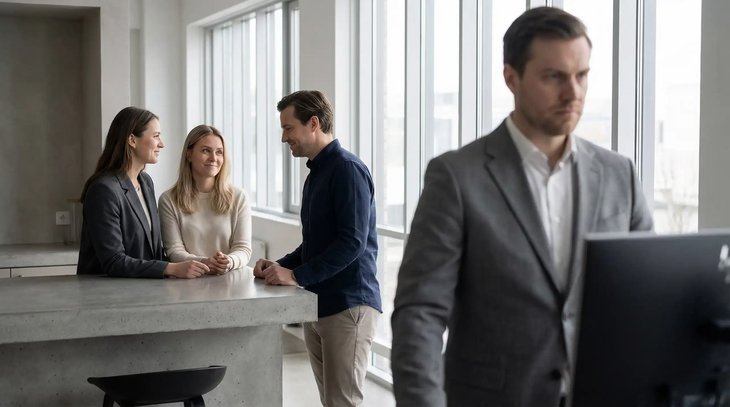 Employé concentré et isolé devant son ordinateur tandis que trois collègues discutent en arrière-plan ; illustration de la surcharge de travail et de l’isolement professionnel.